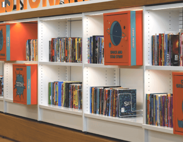 background image: White shelves with books. Some of the shelves have orange boxes within them and have titles describing the themes of the books, like outer space.