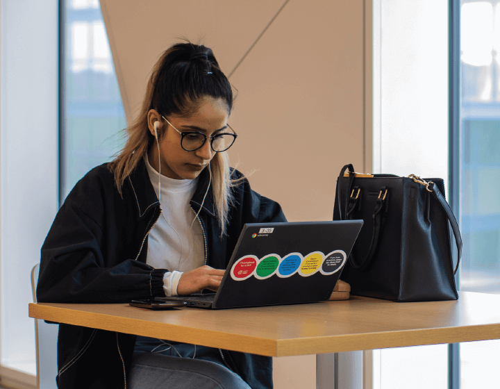 background image: A young woman sits at a table using a black laptop.
