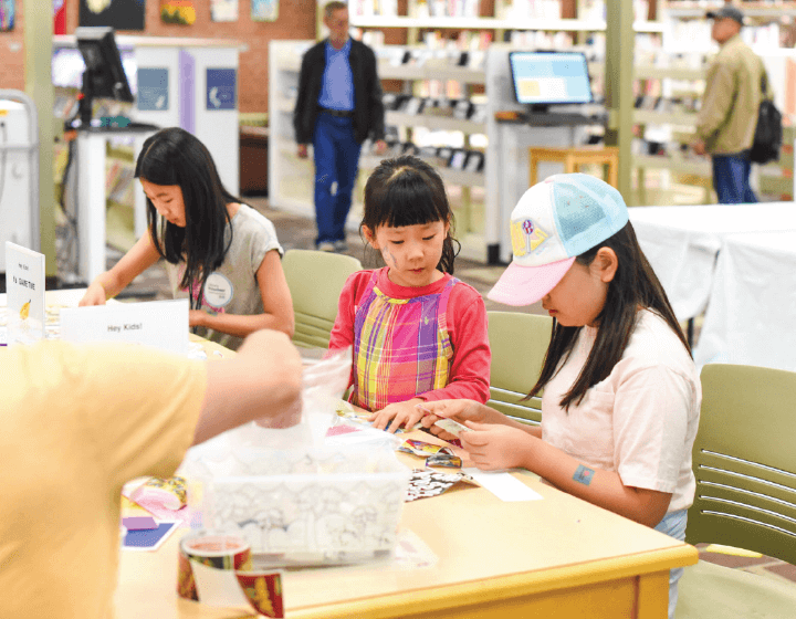 background image: Three children are seated at a table in a library or classroom setting, engaging in a craft activity. They are focused on cutting or assembling materials in front of them. There are various crafting supplies on the table, and other people are visible in t