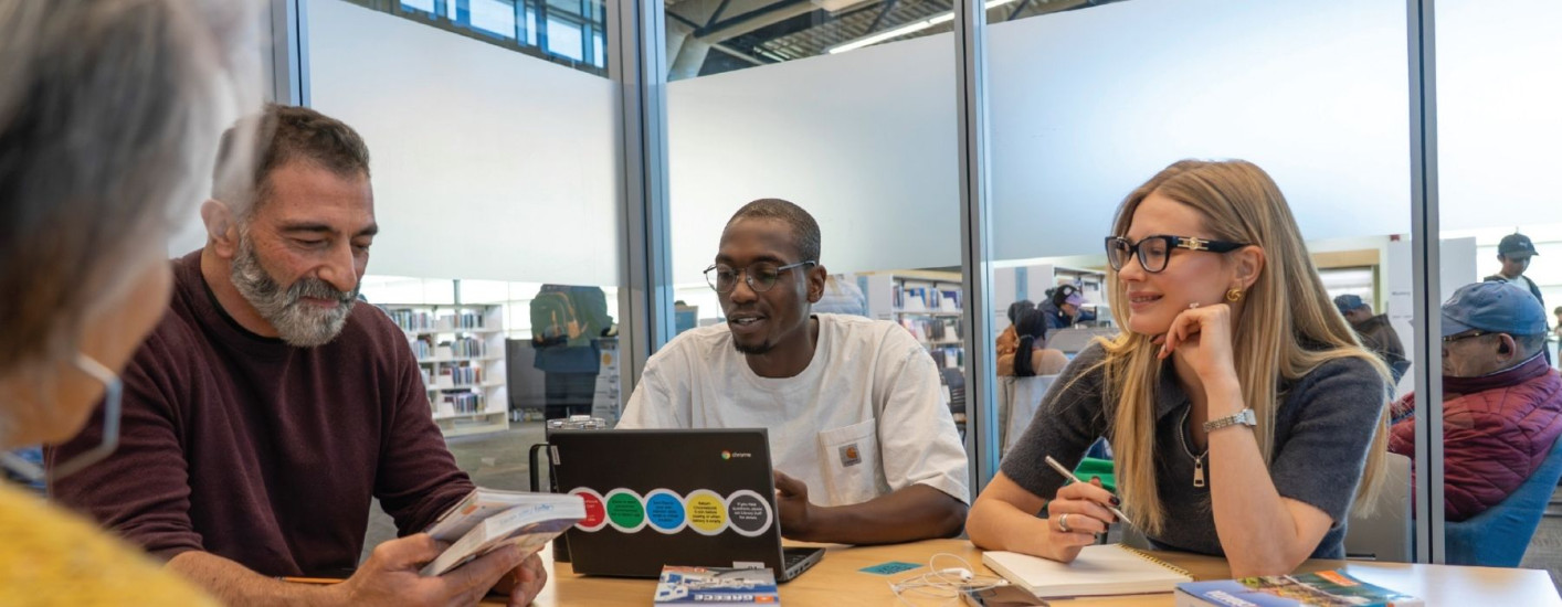 A group of adults sit around a table in a library, collaborating with books and a laptop open in front of them.