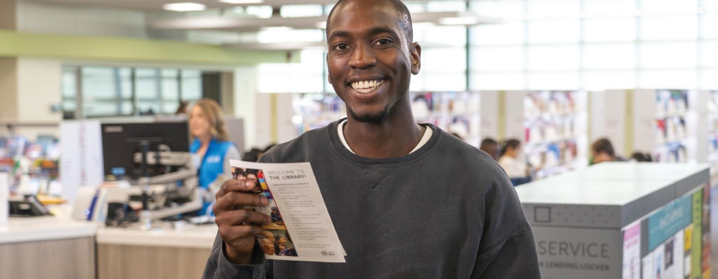 A smiling person holds a library brochure while standing near the service desk at a library.