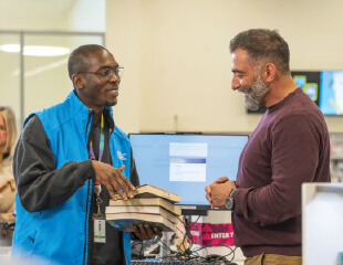 A library staff member hands a stack of books to a smiling visitor at the service desk.