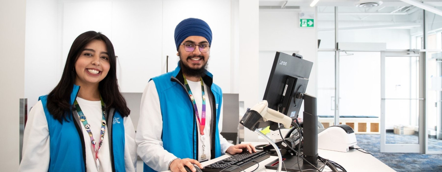 A picture of two Library staff members, a female and a male, standing behind a desk and smiling at the camera.
